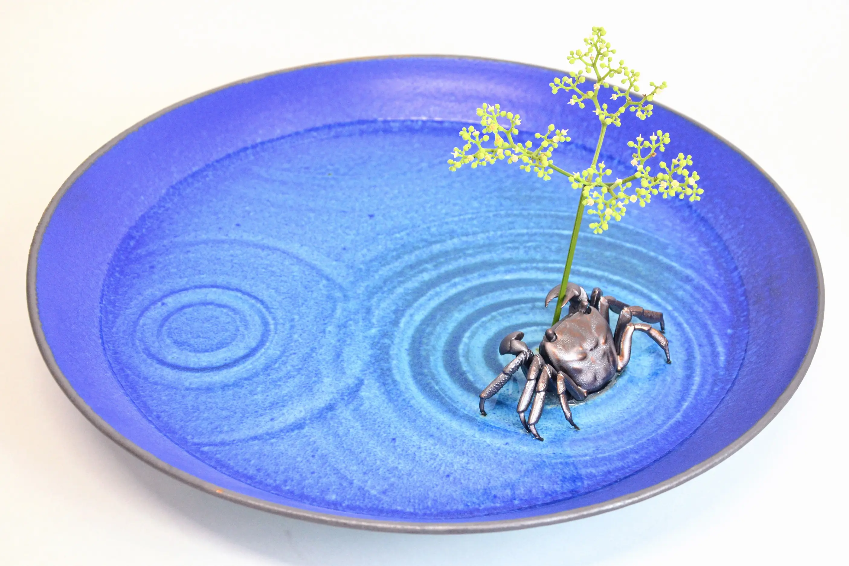 野村俊介の陶芸作品、鮮やかな青色の大皿の上に、小柄な大きさのカニが載っている、大皿はまるで水面のように見えるが、３箇所から波紋が広がっている、一番大きな波紋の中心に切り花が生けられており、金属光沢を帯びた褐色のカニが、片方のハサミでその切り花を挟んでいる。
