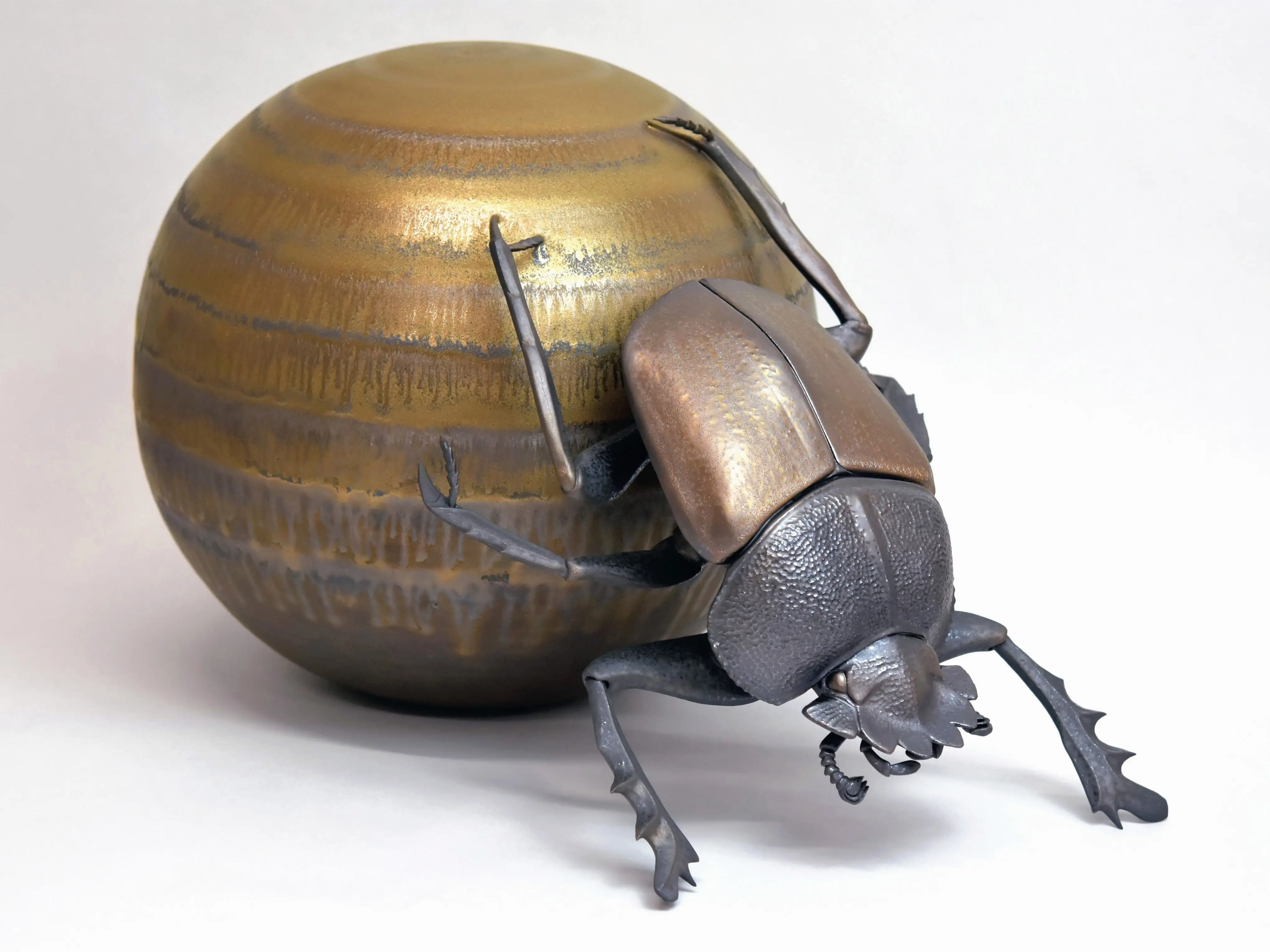 野村俊介の陶芸作品、一匹のスカラベが後ろ足で、金属光沢を有する黄色の球体を転がしている、スカラベは明るめの茶褐色をしており、鈍い光沢を放っている、球体は上半分の方が光沢が強い。