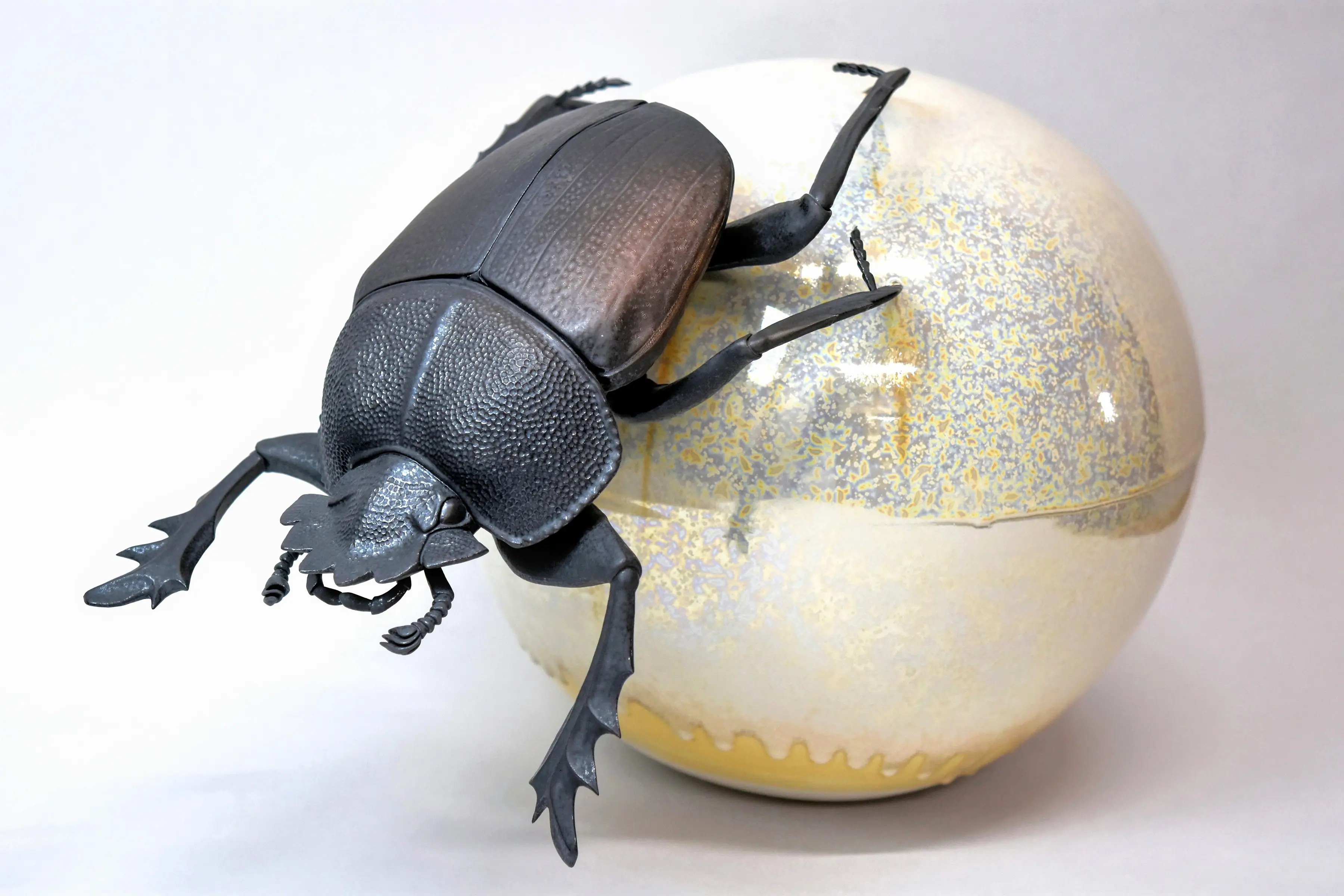 野村俊介の陶芸作品、一匹のスカラベが後ろ足で、光沢のある白色の球体を転がしている、スカラベは鈍く光る茶褐色をしている。