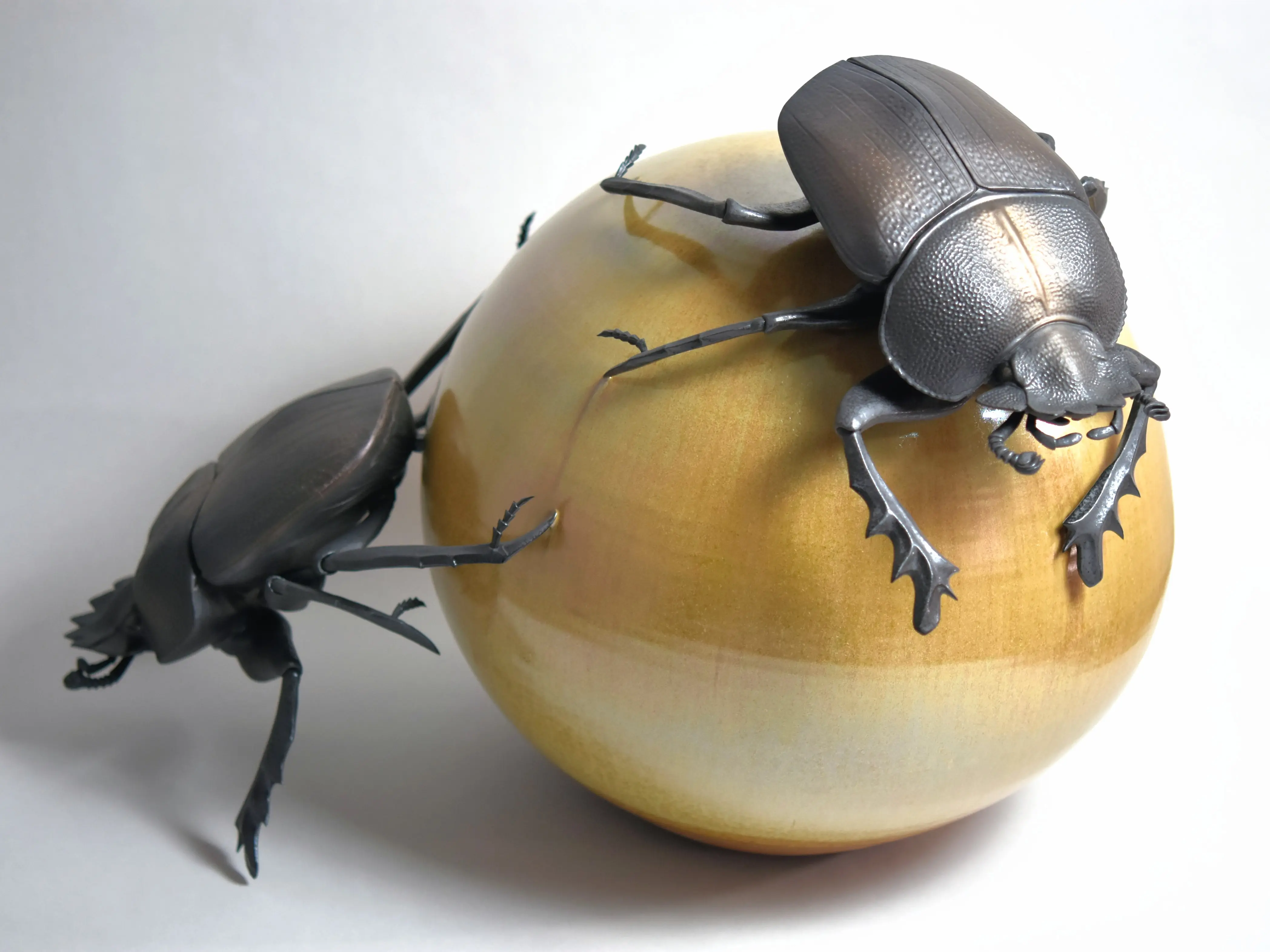 野村俊介の陶芸作品、一匹のスカラベが後ろ足で、黄褐色の球体を転がしている、球体の上にもう一匹のスカラベが載っている、スカラベは鈍く光る茶褐色をしている。